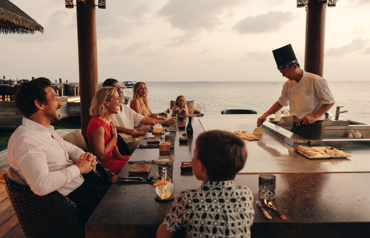 A family laughing together around a waterside restaurant counter at sunset