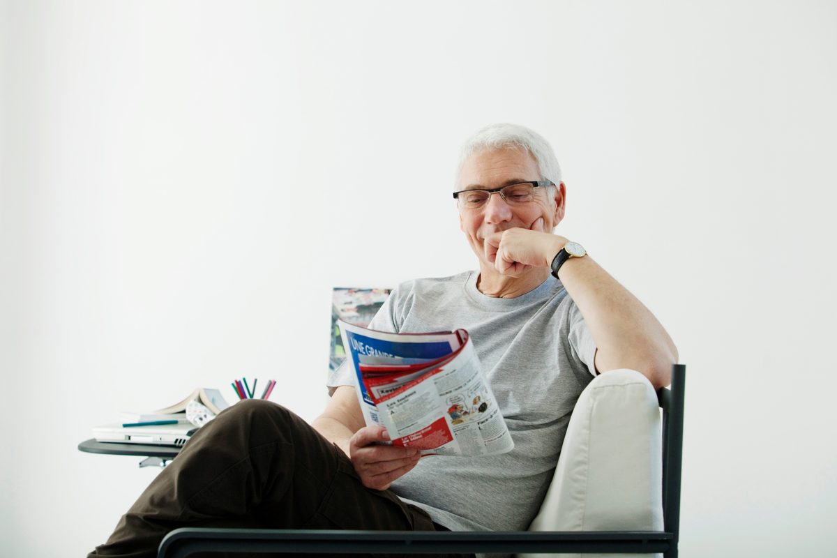 An older man smiling as he reads a magazine in an armchair