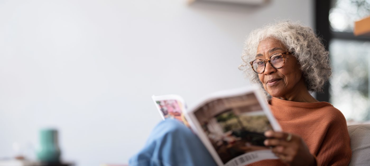 A woman with grey curls smiling as she reads a magazine on the sofa